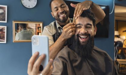 Smiling barber and customer in the barbershop watching a video on a smartphone.