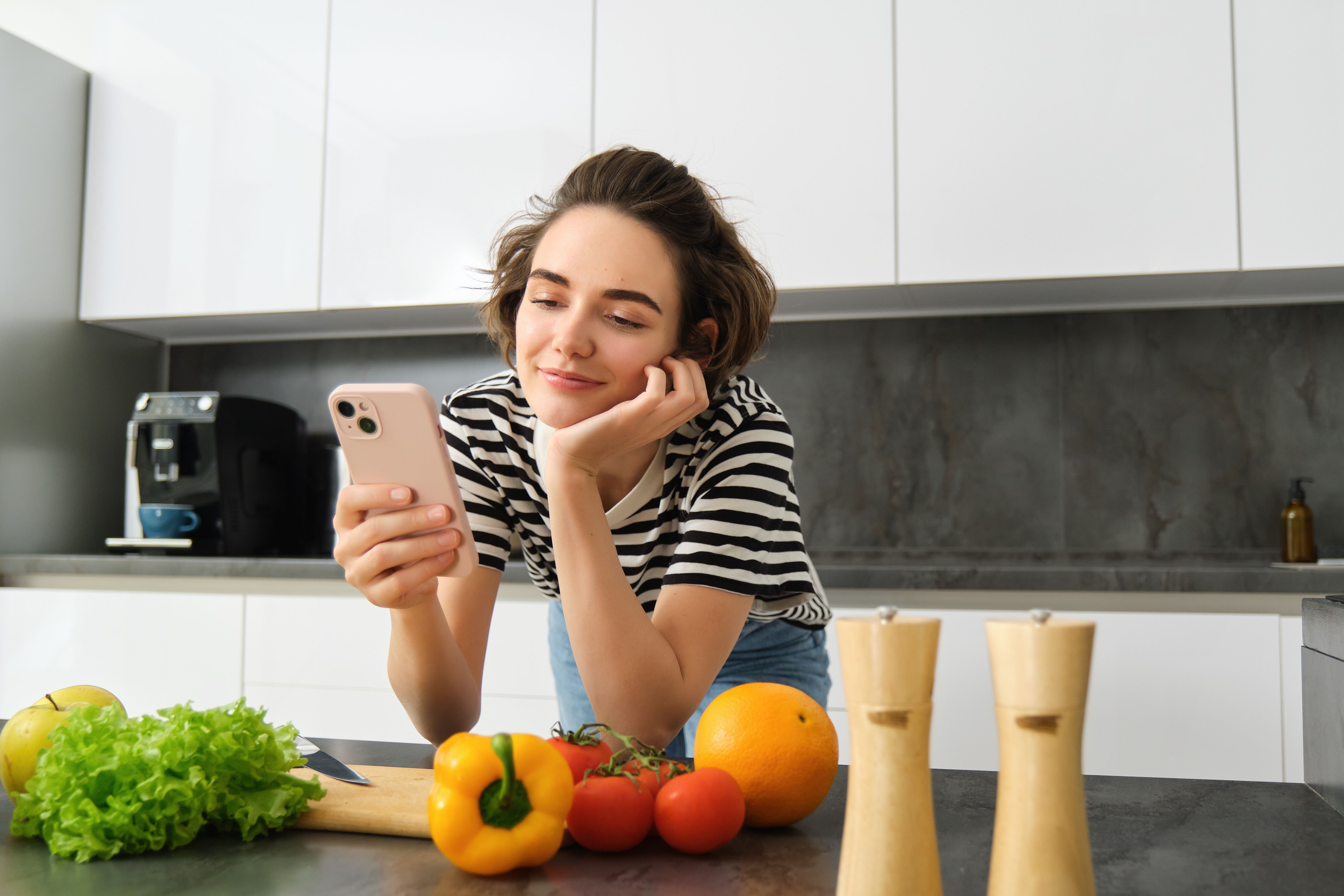 Portrait of beautiful young woman with smartphone, cooking in the kitchen, chopping vegetables, reading recipe online