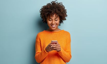 Satisfied hipster girl with Afro haircut, types text message on cell phone, enjoys online communication, types feedback, wears orange jumper, isolated on blue studio wall. Technology concept