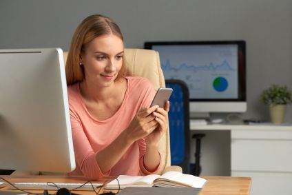 Mulher com blusa rosa claro sentada em uma cadeira de escritório enquanto usa celular para acessar plataforma de serviços financeiros. Na frente dela há uma mesa com caderno aberto e computador.
