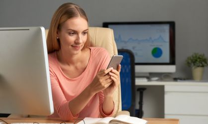 Mulher com blusa rosa claro sentada em uma cadeira de escritório enquanto usa celular para acessar plataforma de serviços financeiros. Na frente dela há uma mesa com caderno aberto e computador.