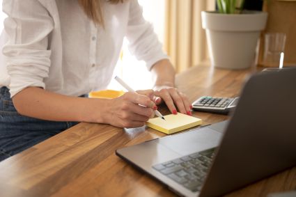 Mulher de camisa branca escevendo em bloco de anotações, com calculadora ao seu lado e notebook à sua frente enquanto utiliza sua conta digital.