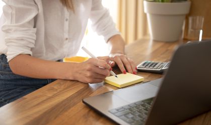 Mulher de camisa branca escevendo em bloco de anotações, com calculadora ao seu lado e notebook à sua frente enquanto utiliza sua conta digital.