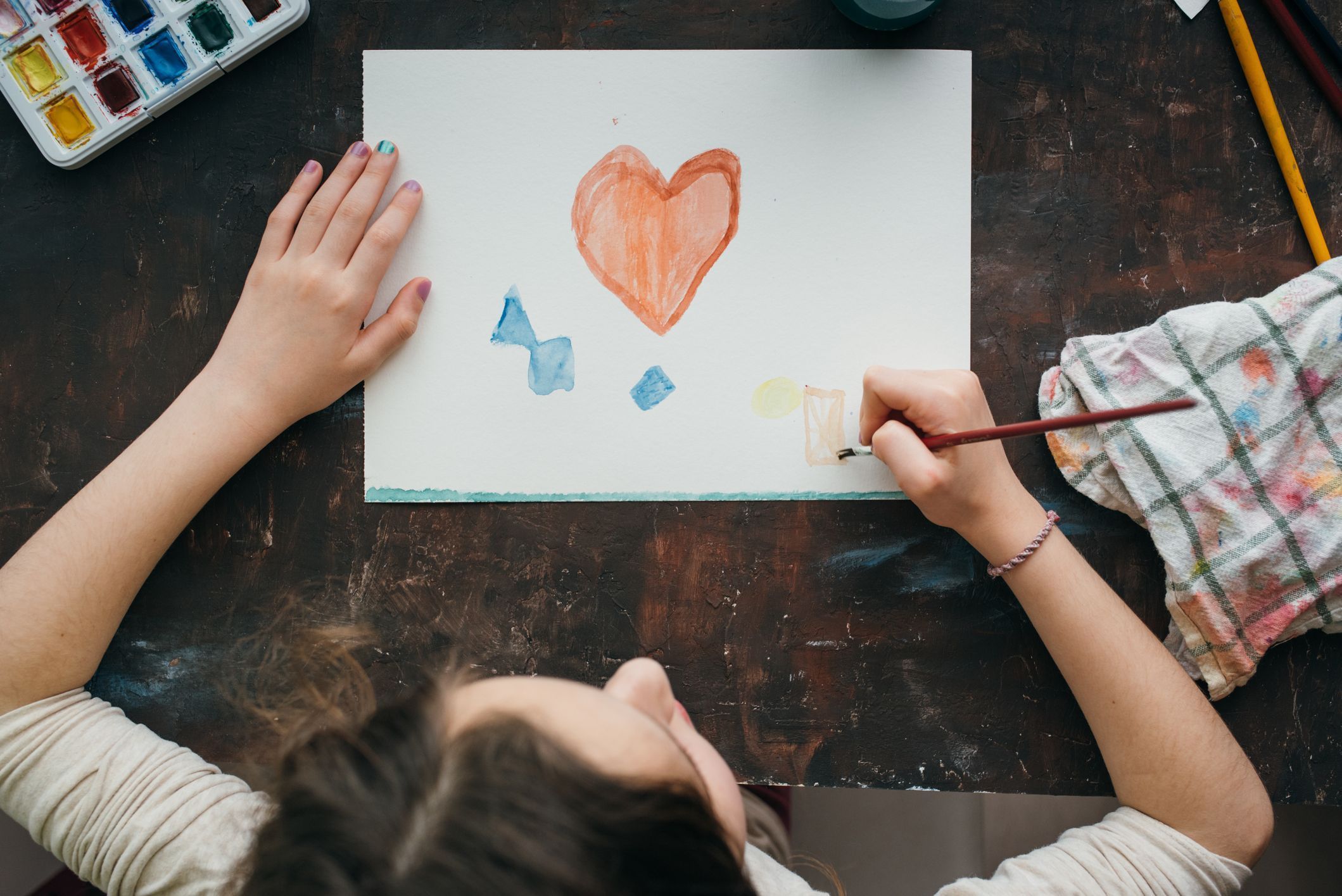 Top view of a girl painting a heart with watercolors on a paper.
