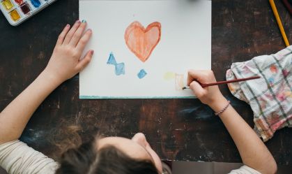 Top view of a girl painting a heart with watercolors on a paper.