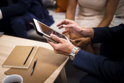Uma pessoa vestida com traje de negócios utiliza um tablet em uma mesa, com um caderno e uma caneta por perto. Outras pessoas são parcialmente visíveis ao fundo.