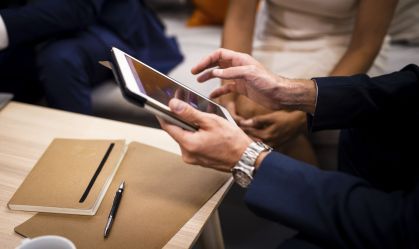 Uma pessoa vestida com traje de negócios utiliza um tablet em uma mesa, com um caderno e uma caneta por perto. Outras pessoas são parcialmente visíveis ao fundo.