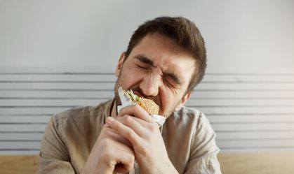 Young unshaven handsome guy with dark hair eating sandwich in fast food with closed eyes, with happy and satisfied expression.