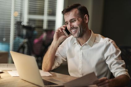 Homeme de camisa branca sorrindo enquanto fala no telefone e olha para tela do notebook que está na frente dele, em cima de uma mesa.
