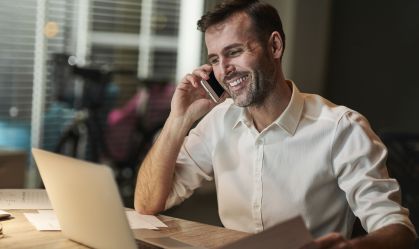 Homeme de camisa branca sorrindo enquanto fala no telefone e olha para tela do notebook que está na frente dele, em cima de uma mesa.