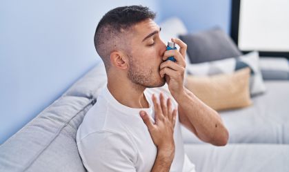 Young hispanic man using inhaler sitting on sofa at home