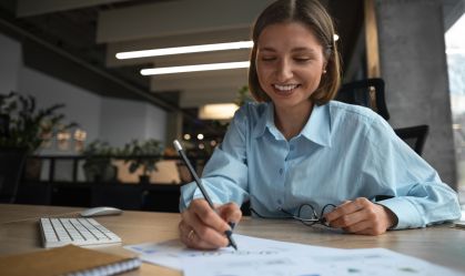 Mulher de camisa azul escreve planejamento financeiro em papéis em cima de uma mesa.