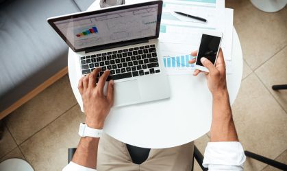 Pessoa utiliza serviços financeiros pelo celular e notebook, com braços apoiados em uma mesa branca redonda.