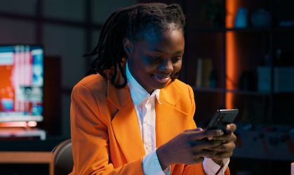 Mulher com blazer laranja sorri enquanto utiliza serviços financeiros com a ajuda de agente de inteligência artificial pelo celular, que segura com as duas mãos.