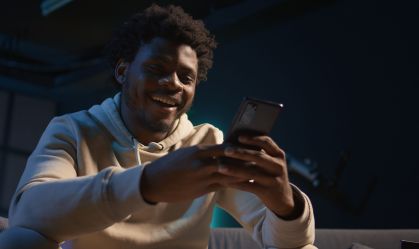 Homem com casaco de moletom sorrindo enquanto digita em celular com as duas mãos.