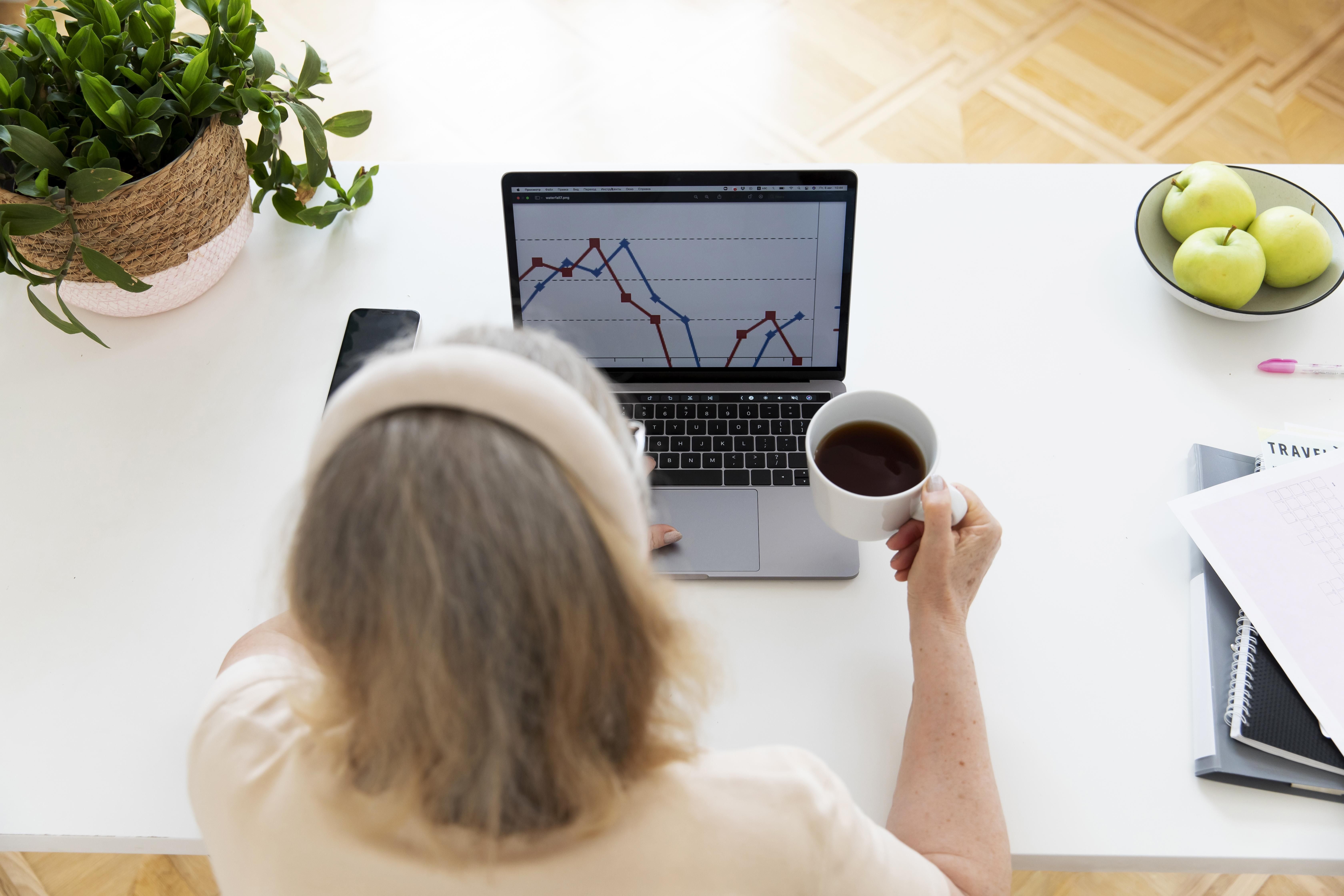 Mulher acompanha gráficos de desempenho dos seus investimentos pelo notebook enquanto toma uma xícara de café.