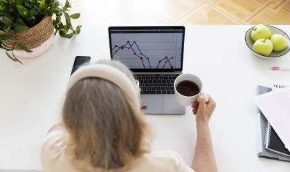 Mulher acompanha gráficos de desempenho dos seus investimentos pelo notebook enquanto toma uma xícara de café.