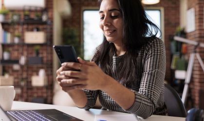 Mulher sorri enquanto usa o celular. Tem os cotovelos apoiados em uma mesa, e um notebook e uma caneta na frente dela.