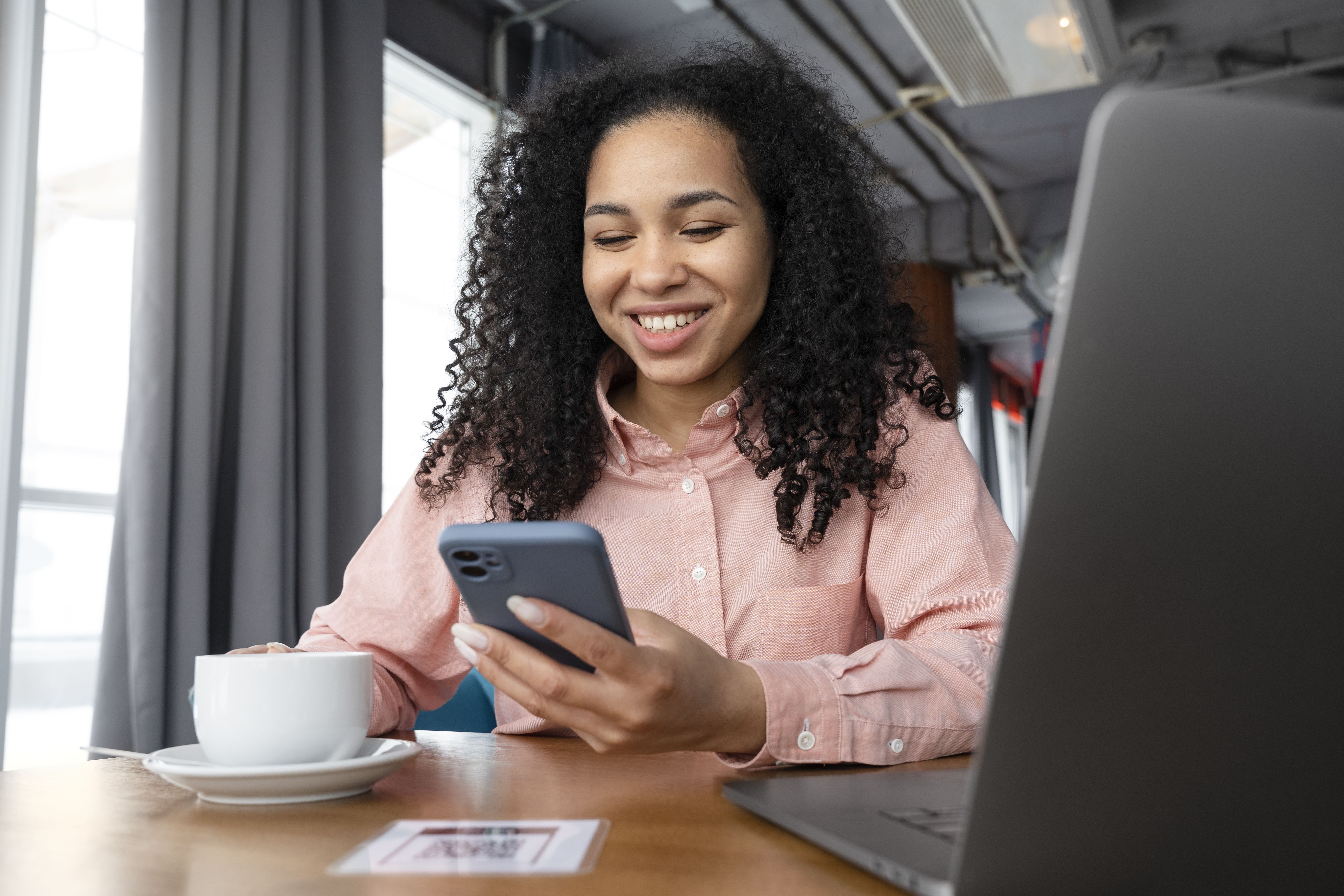 Mulher com camisa rosa realiza transações pelo celular com o braço apoiado em uma mesa que tem uma xícara de café e um notebook.