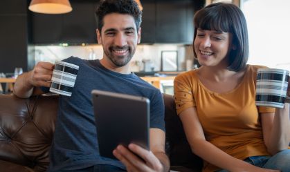 Um homem de camisa azul e uma mulher de blusa amarela, sentados lado a lado, sorriem enquanto usam um tablet para acessar serviços financeiros.