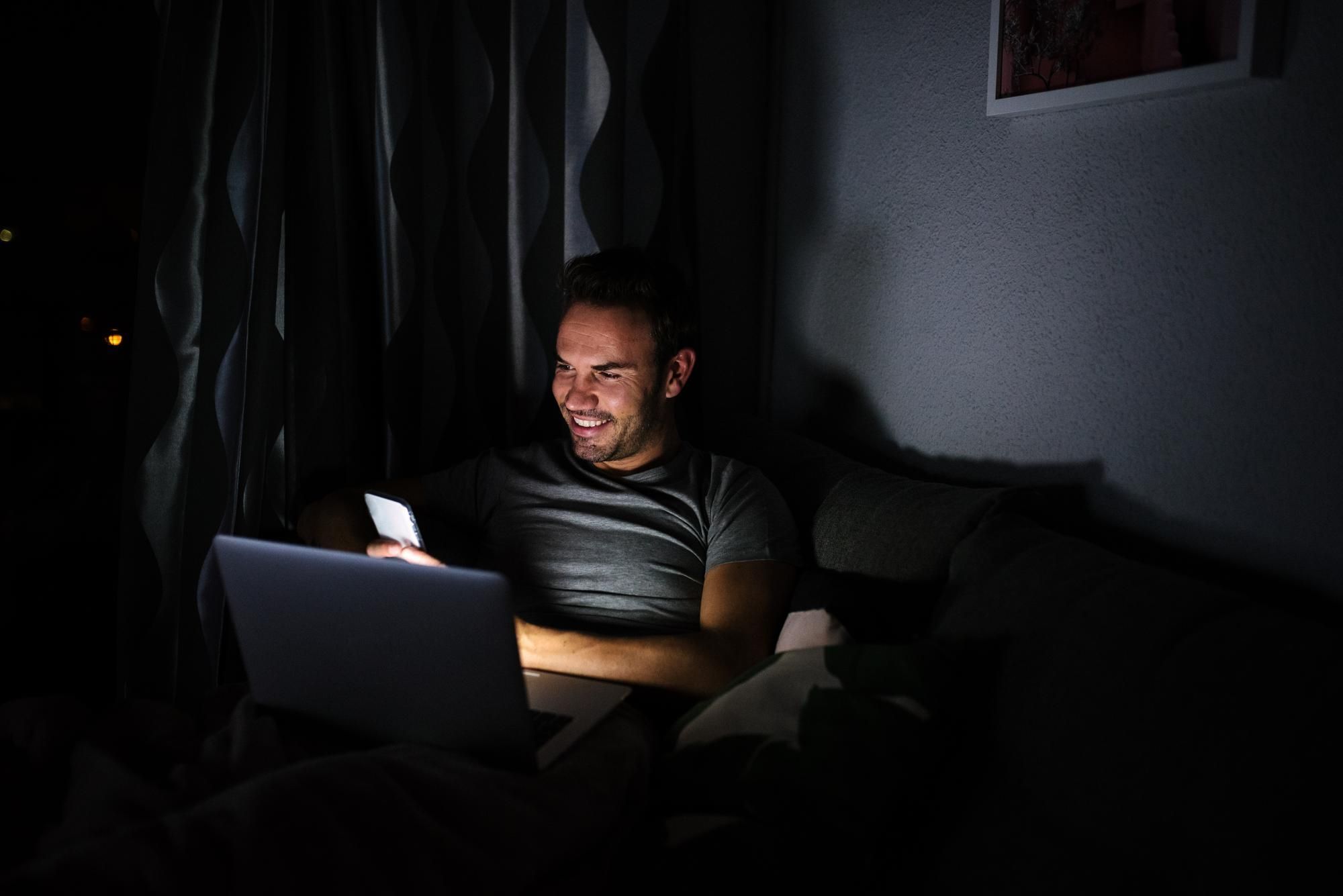 homem-sentado-no-sofá-à-noite-olhando-para-celular-e-computador