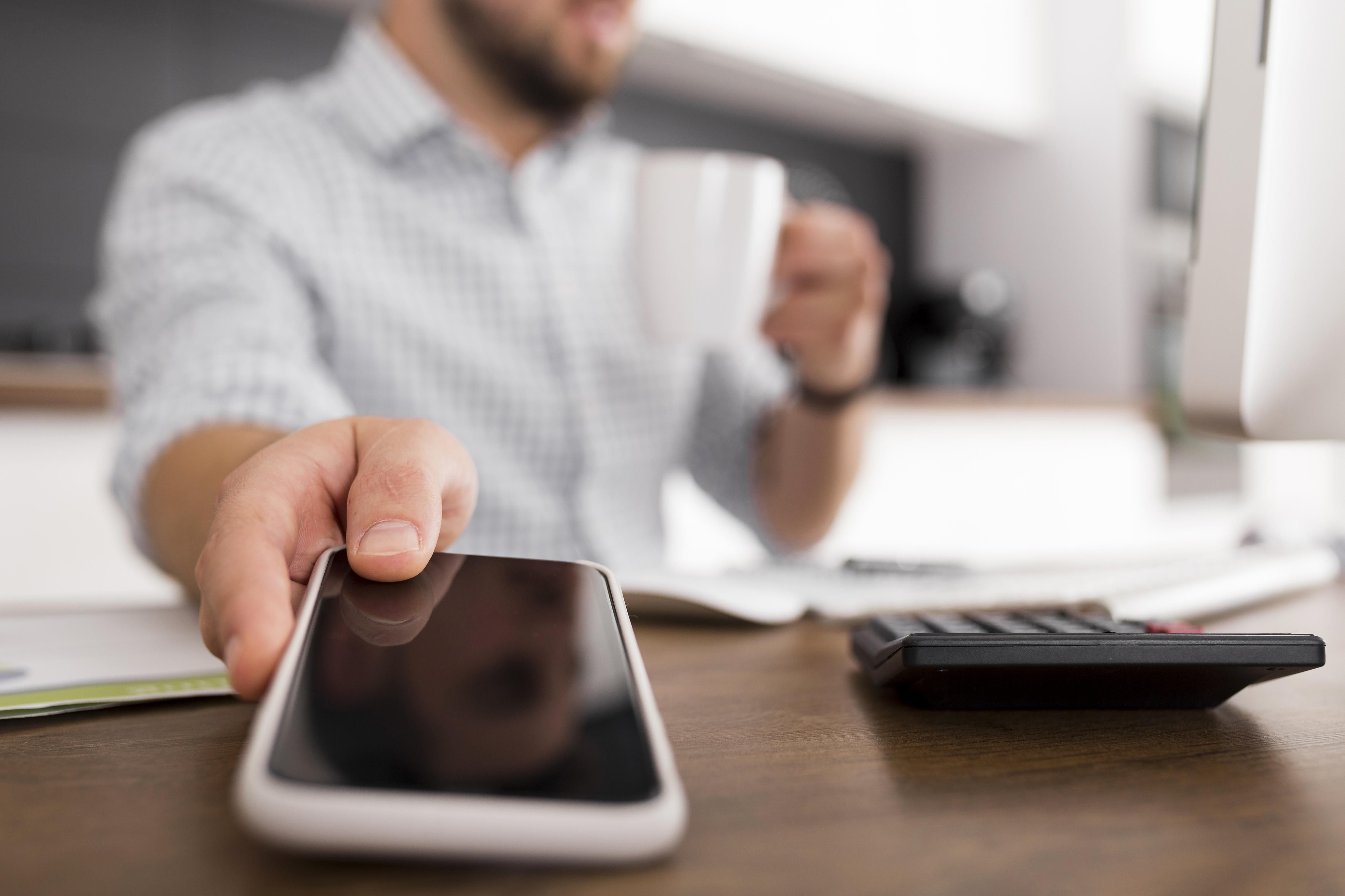 Homem de camiseta branca segura celular em uma mão e xícara na outra, com notebook e papéis à sua frente.