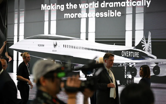 Visitantes passam por um cartaz do conceito de aeronave Overture da Boom Supersonic no Farnborough Airshow, no condado de Hampshire, Inglaterra, em julho de 2022 • Justin Tallis/AFP/Getty Images