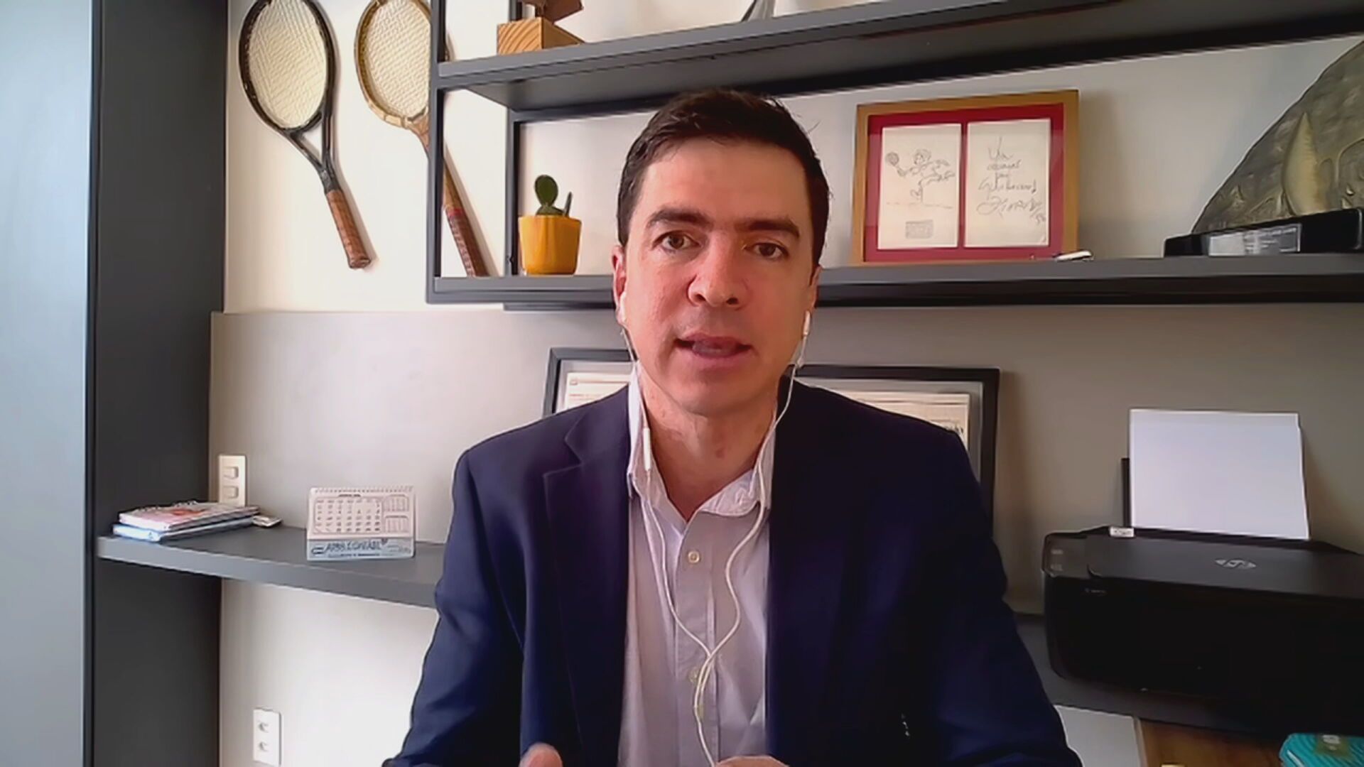 Gabriel Dietze está sentado, vestindo um paletó azul e uma camisa branca. Atrá dele, aparecem prateleiras com itens de decoração, impressora e porta-retratos
