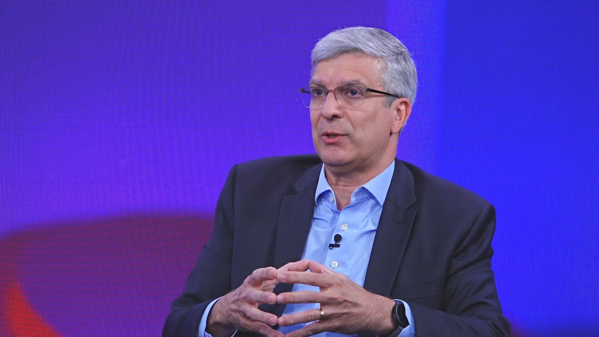 Leando Vilain aparece sentado em uma cadeira, ao fundo de um tellão azul e roxo, durante entrevista do Capital Insights.