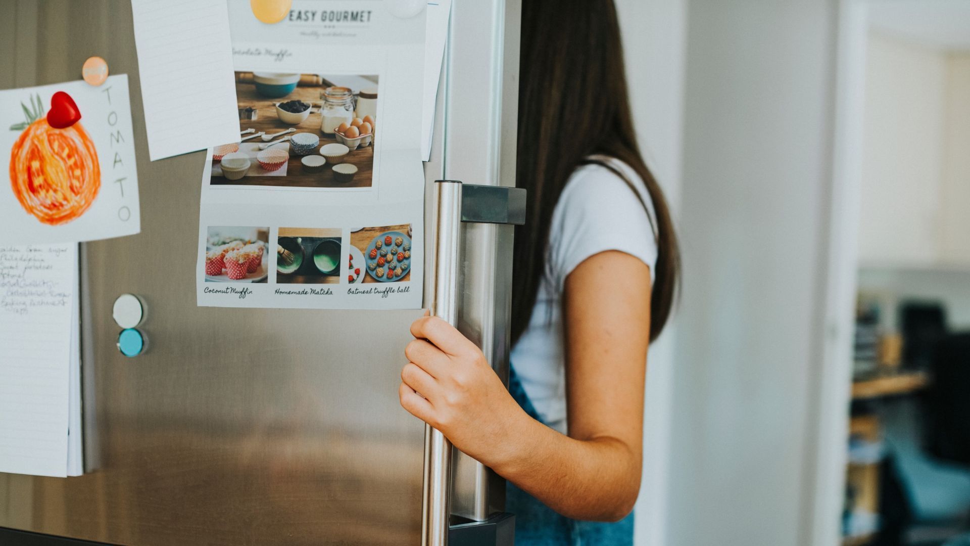 Menina abre uma geladeira decorada com ímãs e fotografias na porta