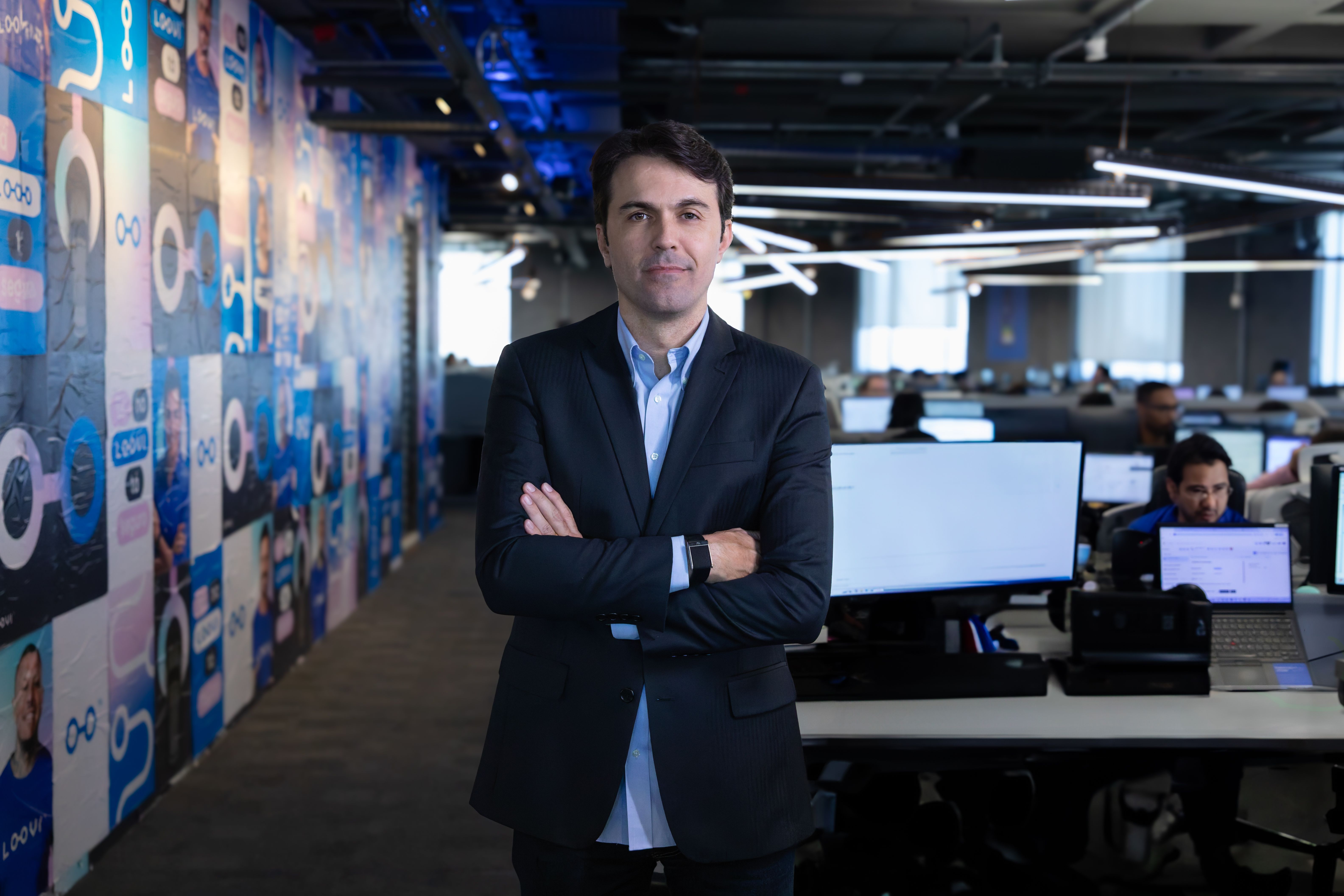 Quezide Cunha, CEO da Loovi, sorrindo e de braços cruzados no escritório corporativo da Loovi, com computadores ao fundo.