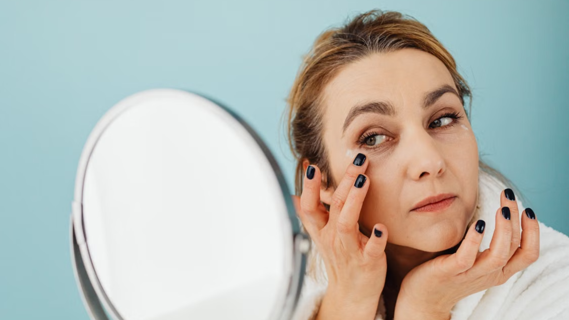 Mulher observa as olheiras diante de um espelho enquanto aplica um produto na região abaixo dos olhos.