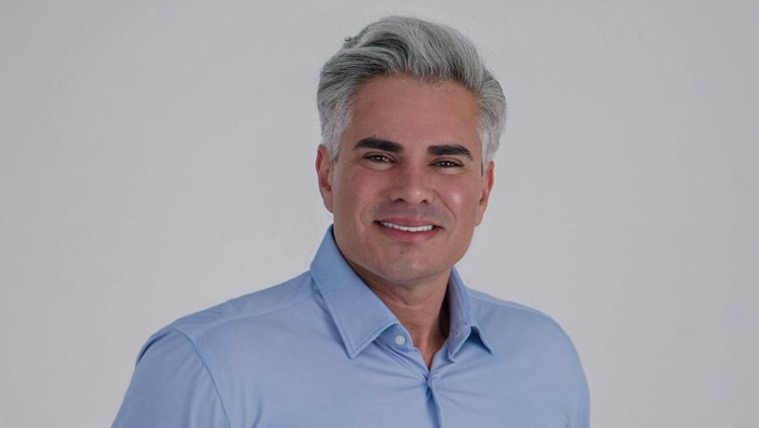 Homem branco com cabelo grisalho veste camisa social azul clara em frente a um fundo neutro