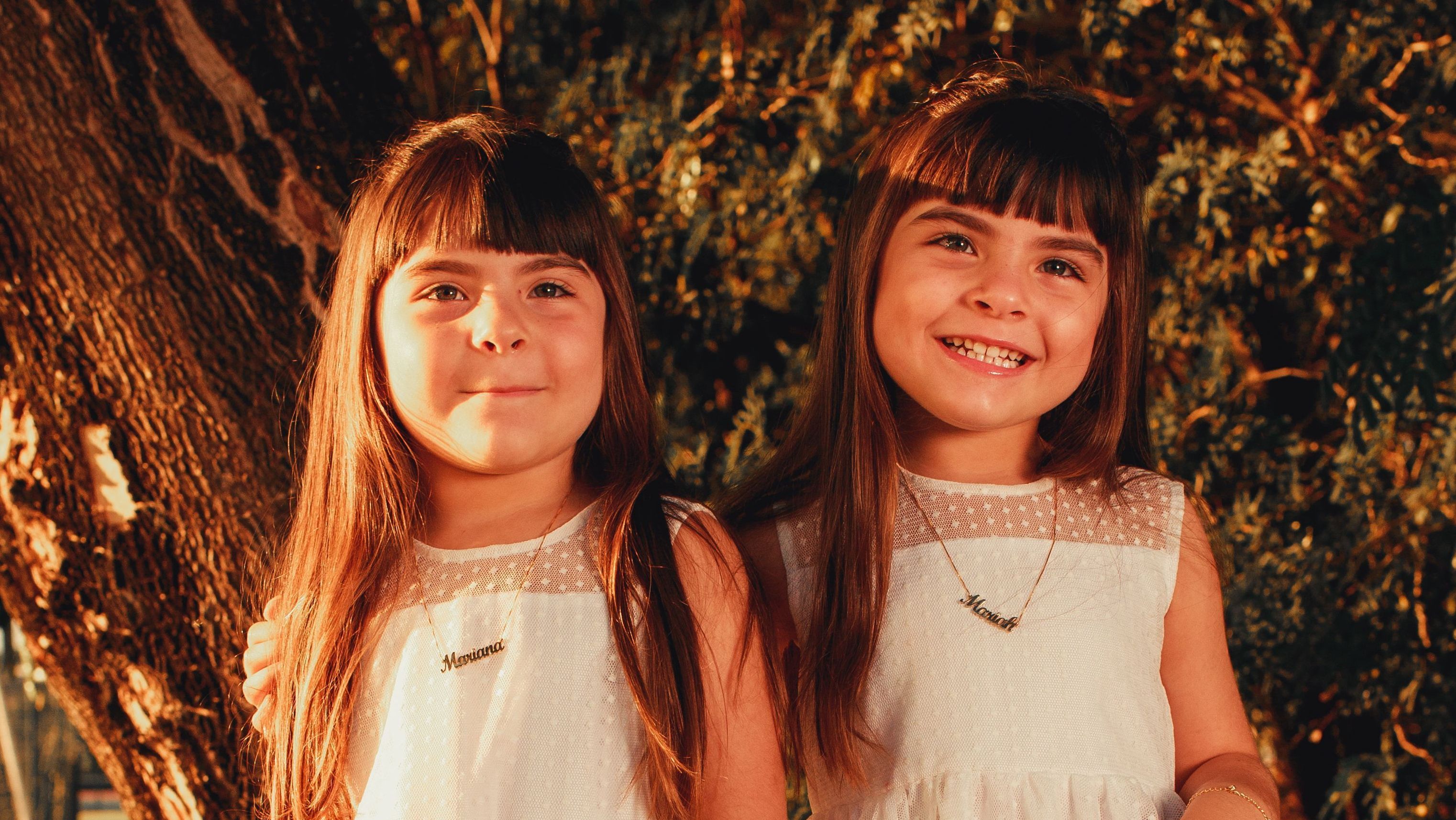 Meninas irmãs gêmeas posam com roupas iguais, cortes de cabelo iguais e colares escrito "Mariah" e "Mariana"