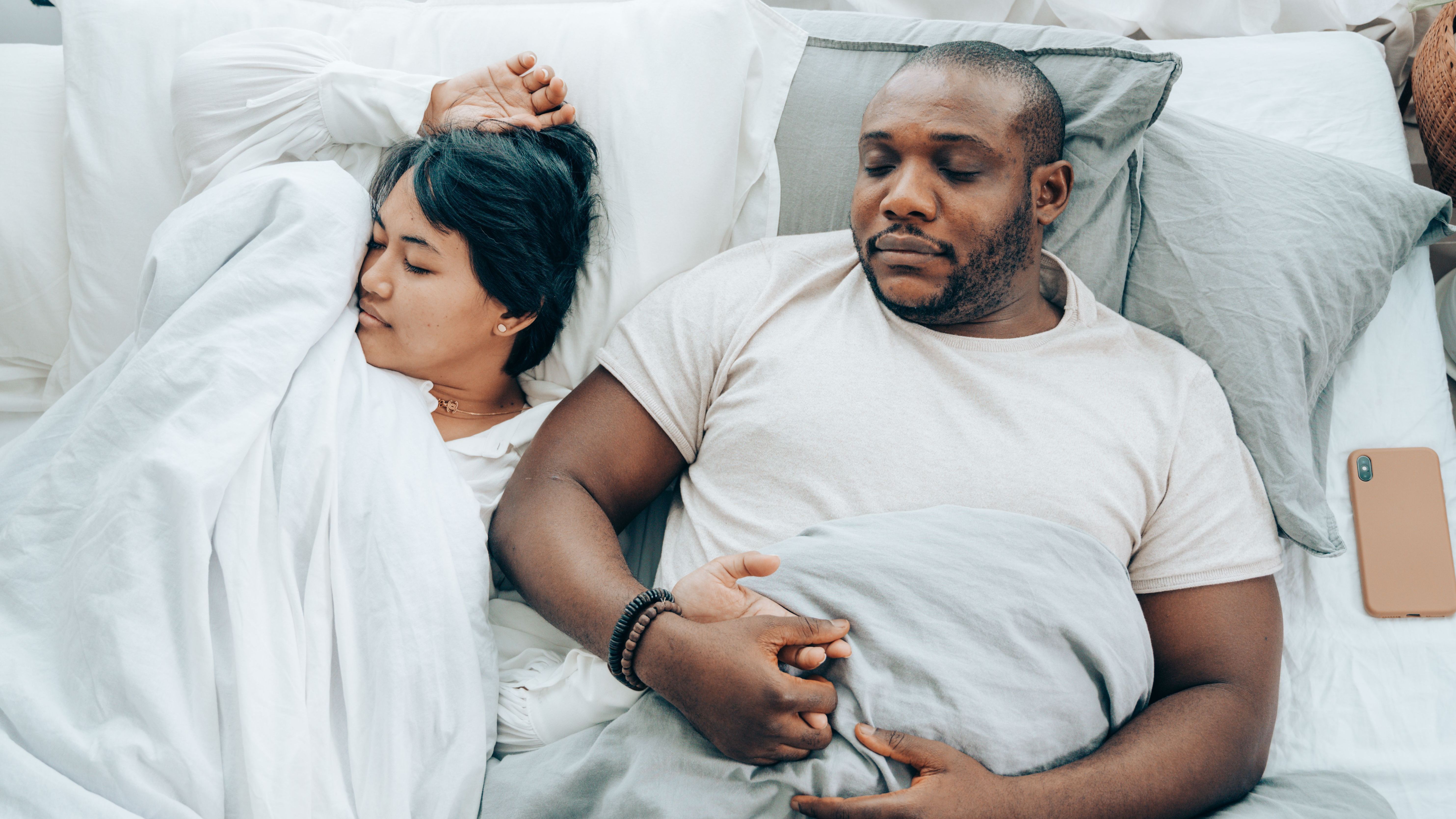 Casal com homem negro e mulher asiática dorme de pijama branco, com coberta, lençóis e fronhas brancas, e celular laranja ao lado