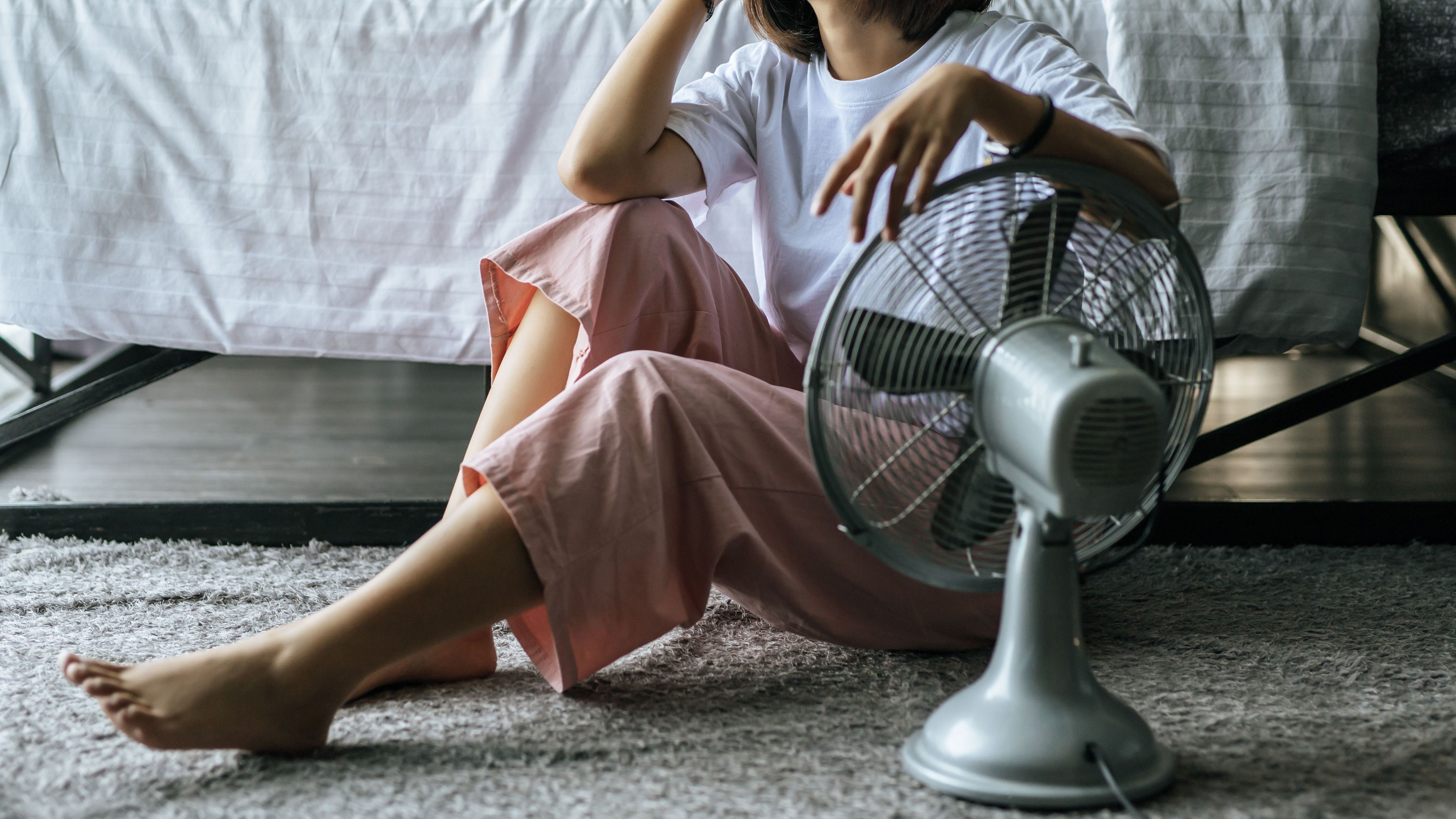 Mulher de camiseta branca e calça rosa, descalça, está sentada ao lado de um ventilador branco, no chão