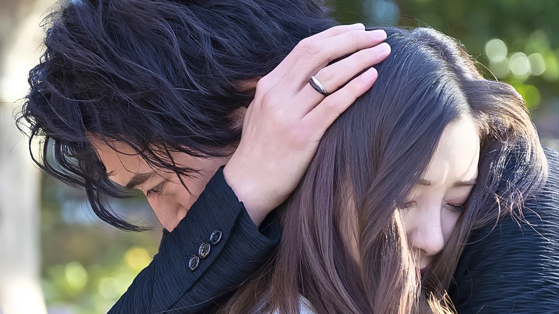 Homem e mulher japoneses se abraçam; a mulher está de olhos fechados, e o homem está a consolando, afagando o cabelo dela