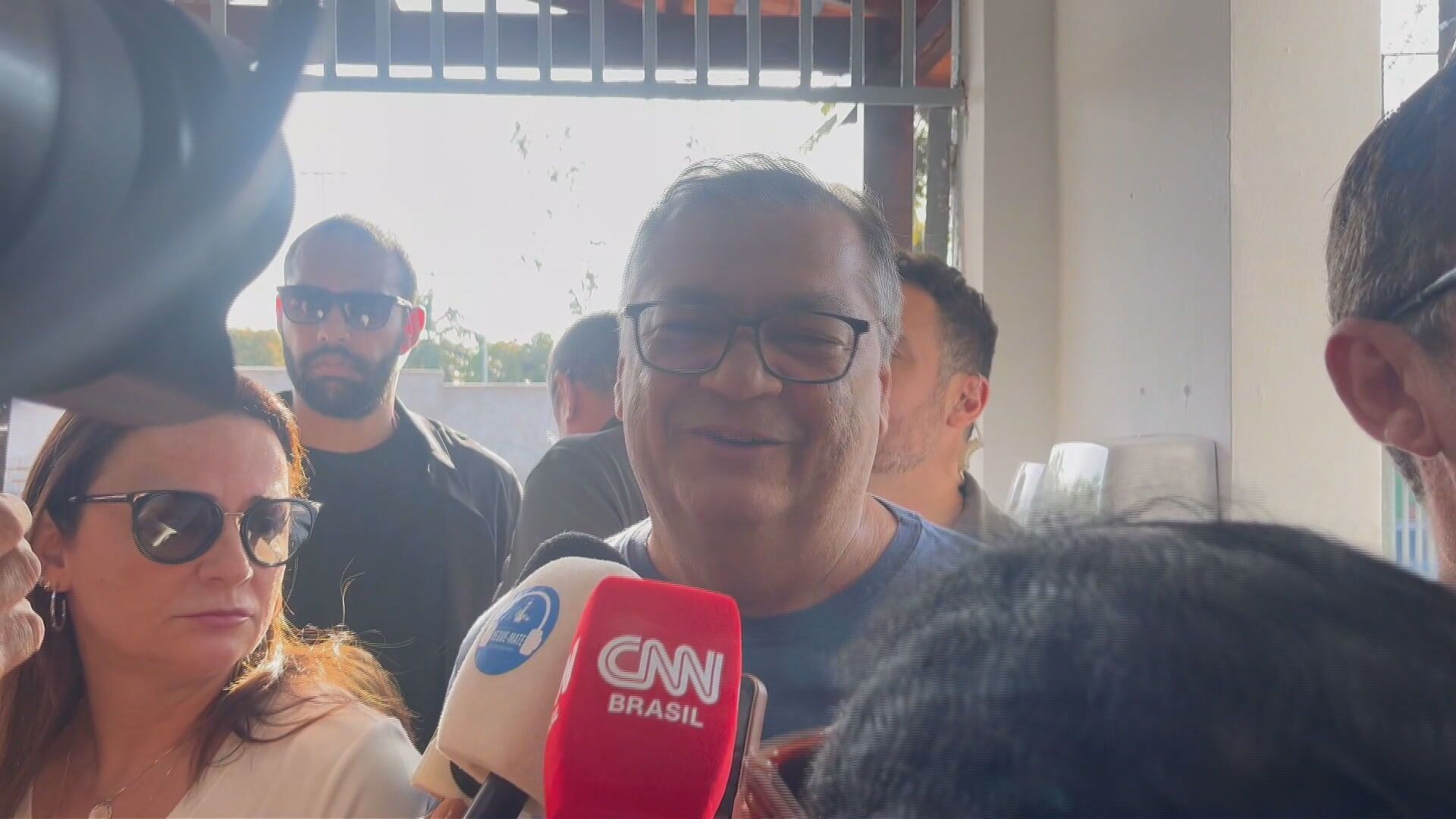 Flávio Dino com óculos e camisa cinza, falando com a imprensa