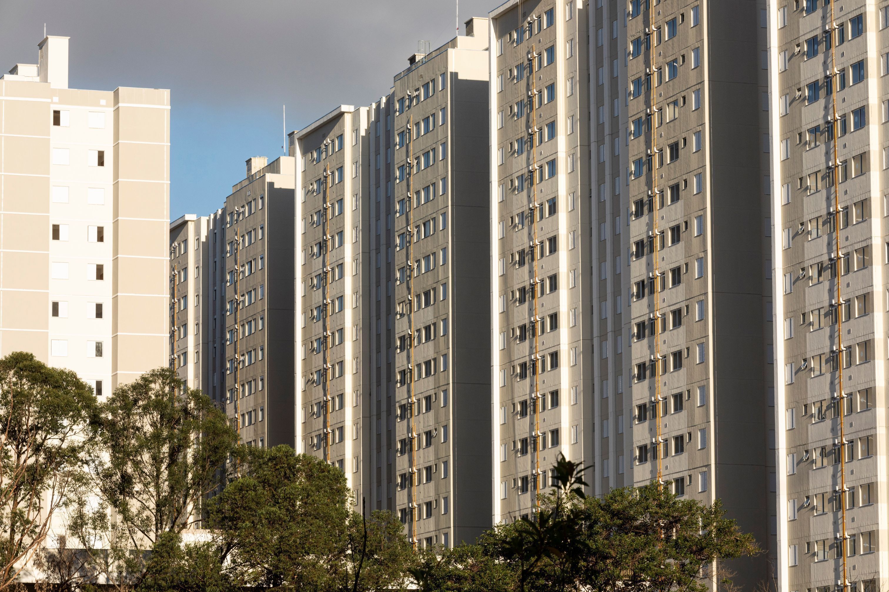 Fila de três prédios de 10 andares nas cores bege e cinza em construção, na parte de abaixo se vê a copa de algumas árvores