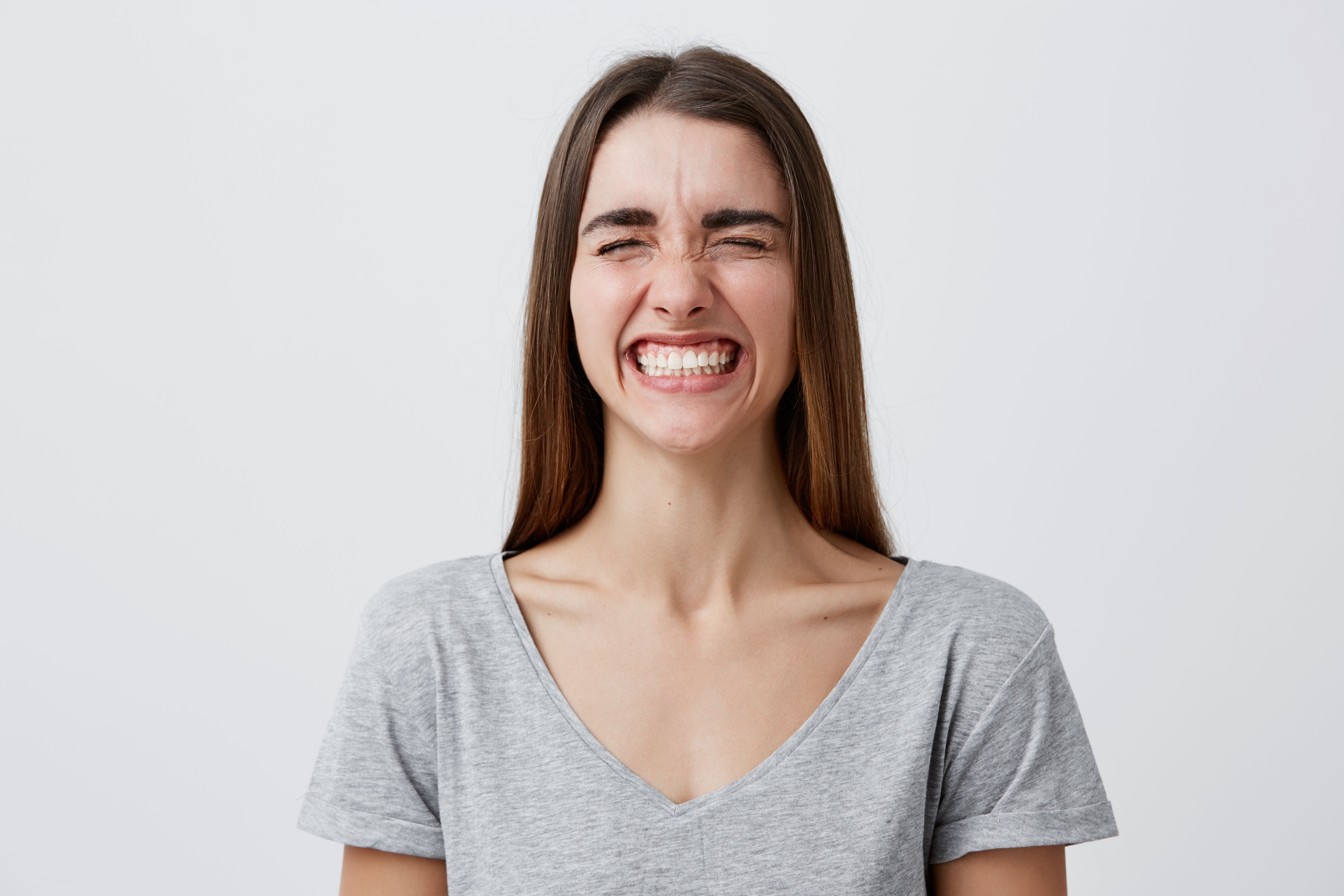 Young attractive cheerful caucasian student girl with dark long hair in casual gray t-shirt laughing brightly wit closed eyes on funny joke of friend in university, being happy and carefree. Positive emotions