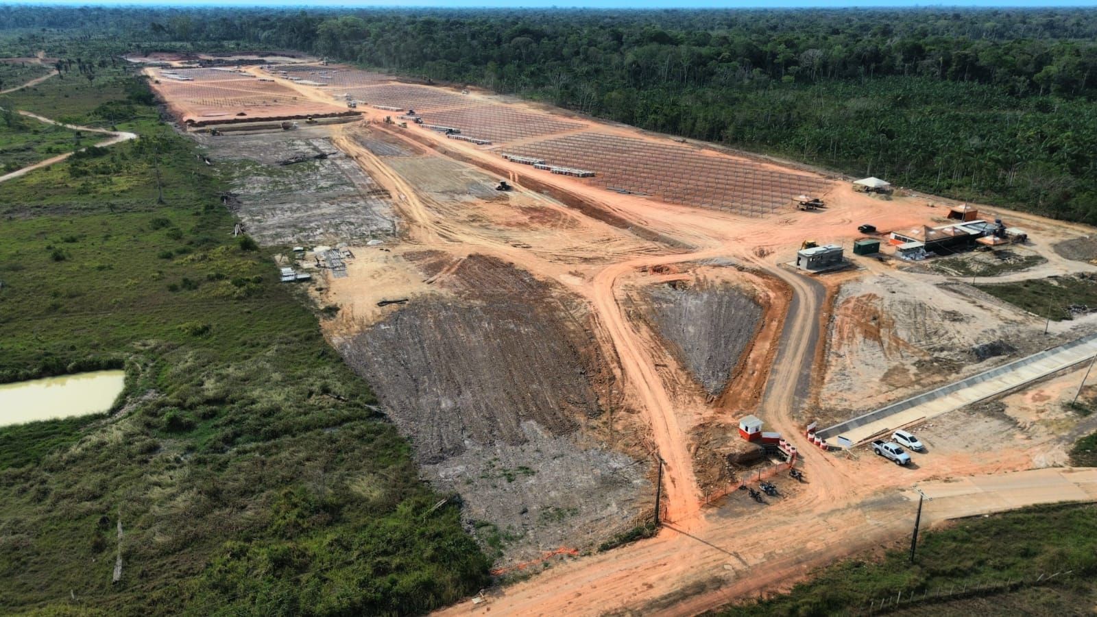 Vista aérea da área em construção da Usina de Codajás, no Amazonas, com terreno nivelado, estruturas metálicas alinhadas, vias de acesso e veículos, cercada por vegetação.