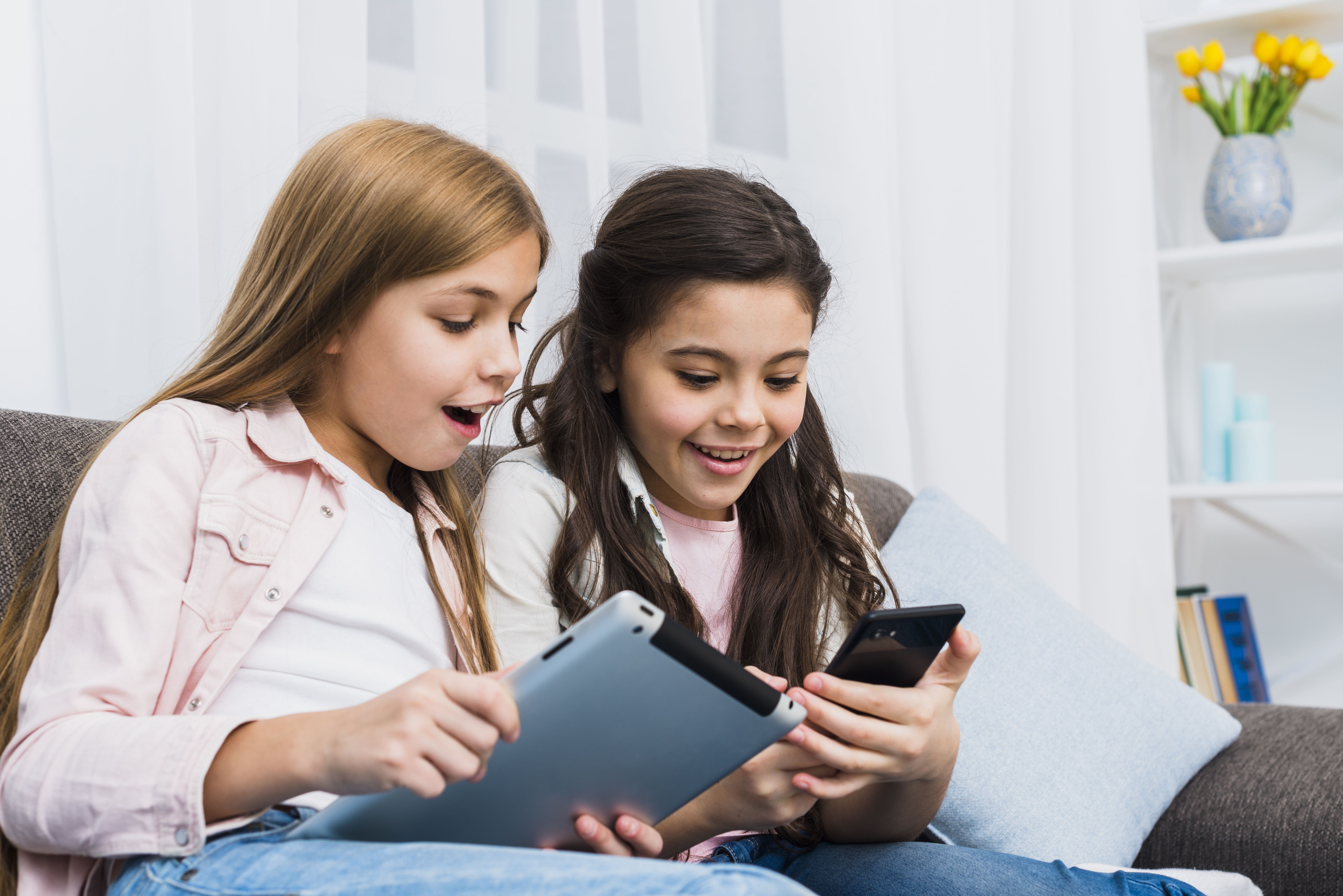 Duas crianças sorriem enquanto olham para o celular e o tablet.