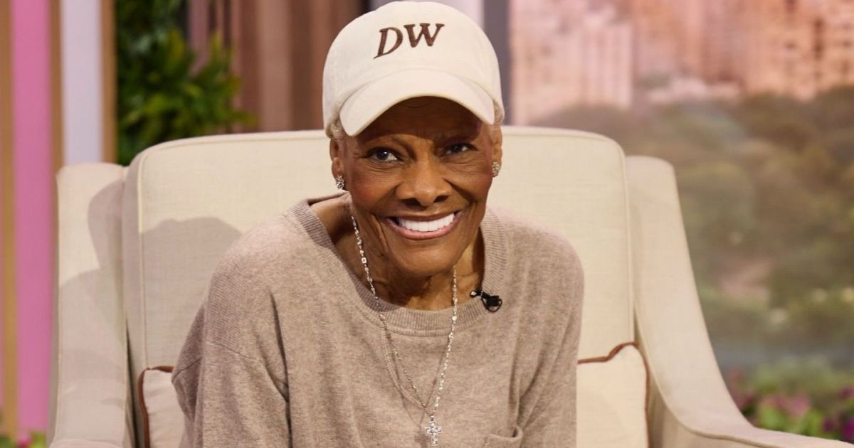 Dionne Warwick em apresentação antiga, sorridente, com microfone na mão.