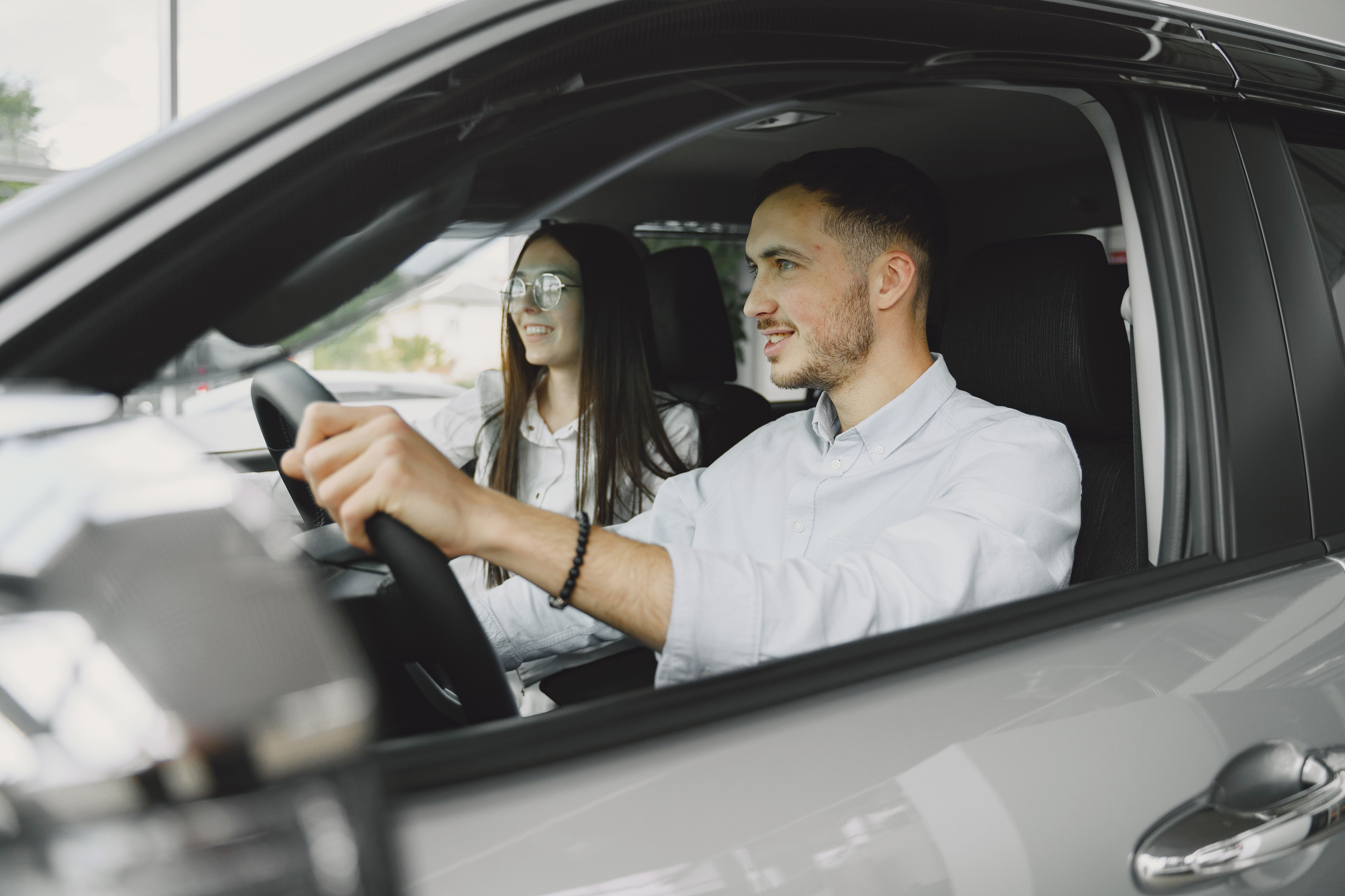 Imagem mostra um carro prateado e dentro dele um casal. O homem está dirigindo com as mãos ao volantes e ao lado uma mulher de cabelos soltos e lisos de óculos. Ambos usam roupas camisas brancas.