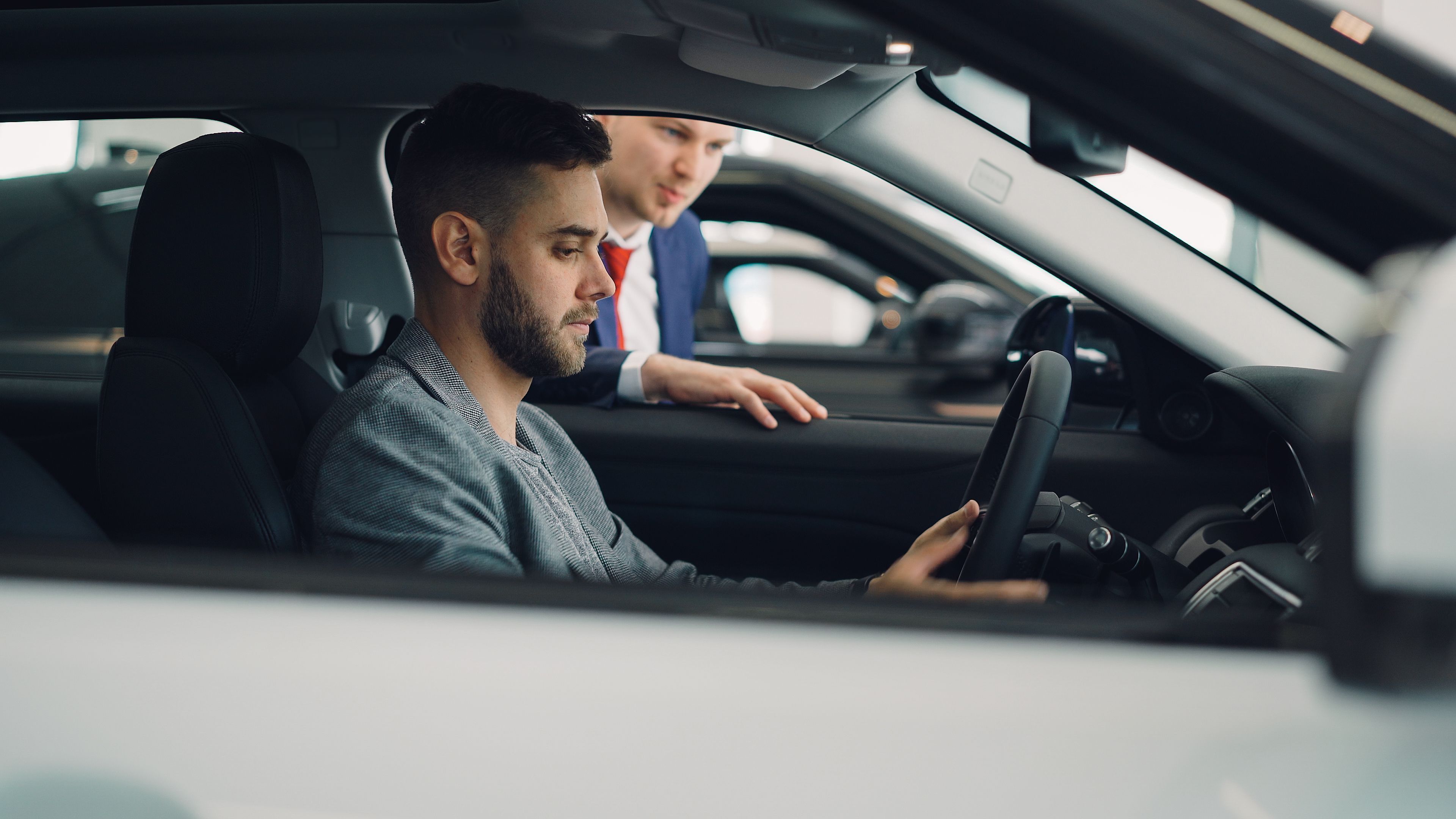 Fotografia mostra um homem branco de perfil, sentado à frente da direção de um carro branco. Do lado de fora, homem branco de terno parece dar instruções