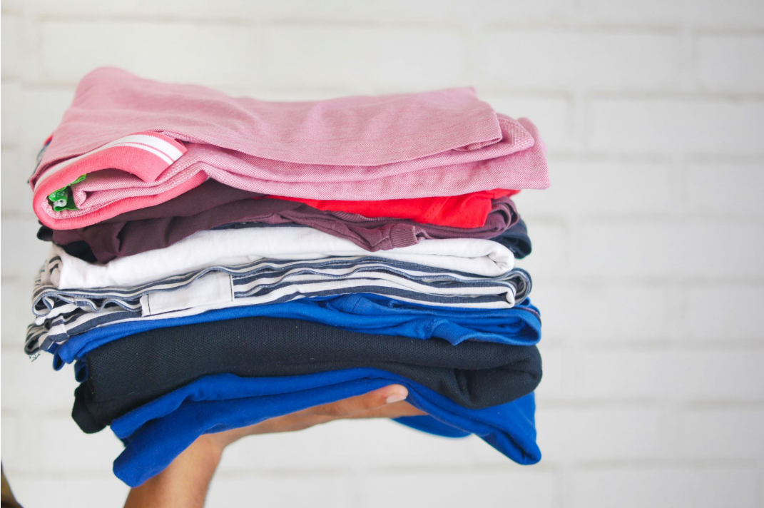 Pessoa segurando uma pilha de roupas dobradas e limpas, em tons de rosa, vinho, branco, listrado, azul e preto, diante de uma parede de tijolos brancos.