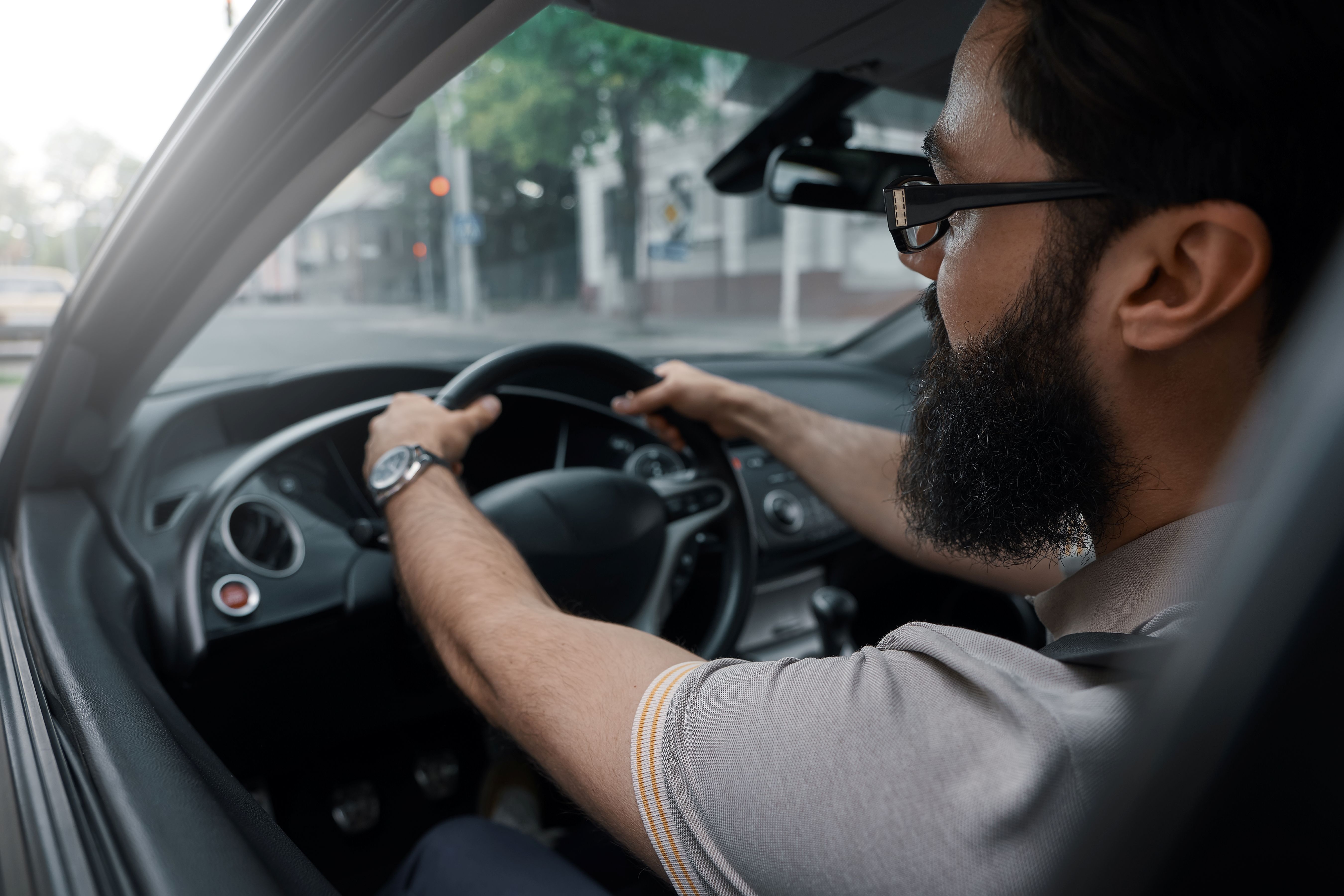 Homem barbudo dirigindo carro, o homem está com óculos de grau e um relógio no pulso esquerdo e está com a mão ao volante