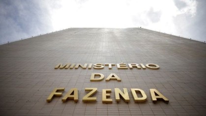Imagem apresentam o pre´dio do Minstério da Fazne,da em uma visão de baixo para cima, mostrando as letras do ministério em dourado e um céu parcilamento nublado